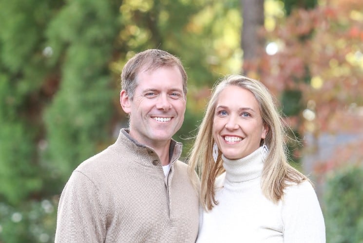 A man in a brown pullover standing with a woman in a white turtleneck outdoors by colorful trees