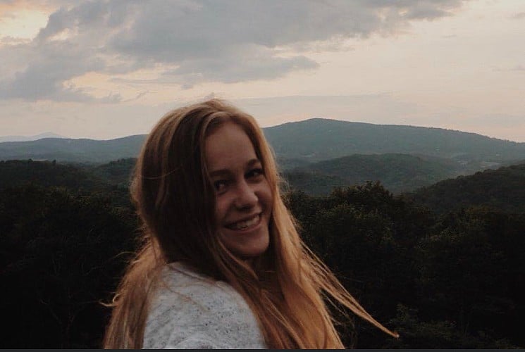 A young girl smiling with an expansive mountain range covered in trees behind her