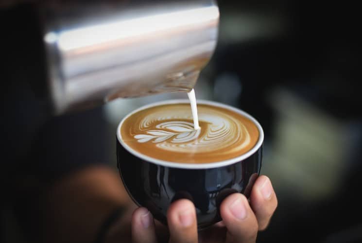 A person holding a black cup of coffee while cream is being poured in to make a decorative design