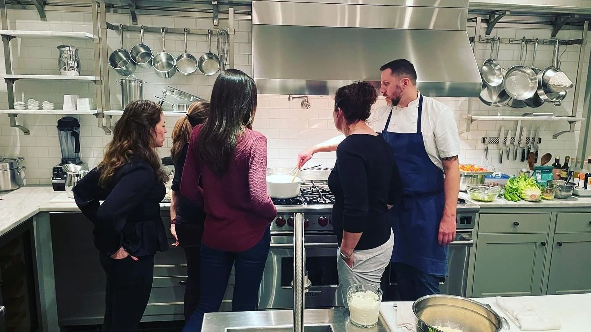 A chef in a blue apron standing at the stove in a chef's kitchen teaching four women