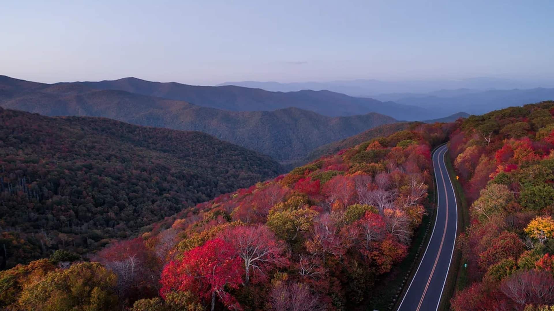 A two-lane road winding within fall colored trees in an expansive mountain range with a hazy sky
