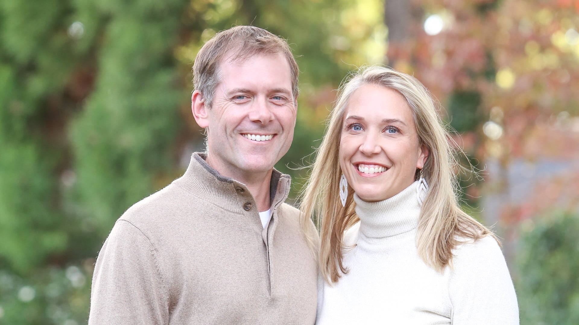 A man in a brown pullover standing with a woman in a white turtleneck outdoors by colorful trees