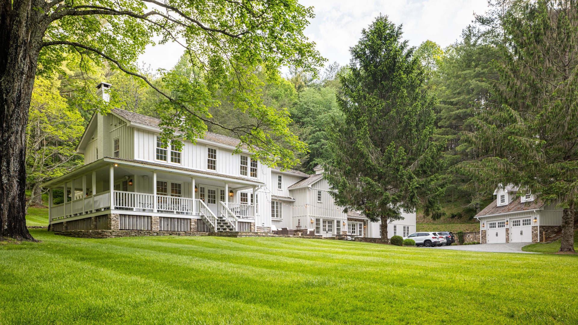 A large white two-story home with wrap around porch, expansive lawn, tall trees and detached double stall garage