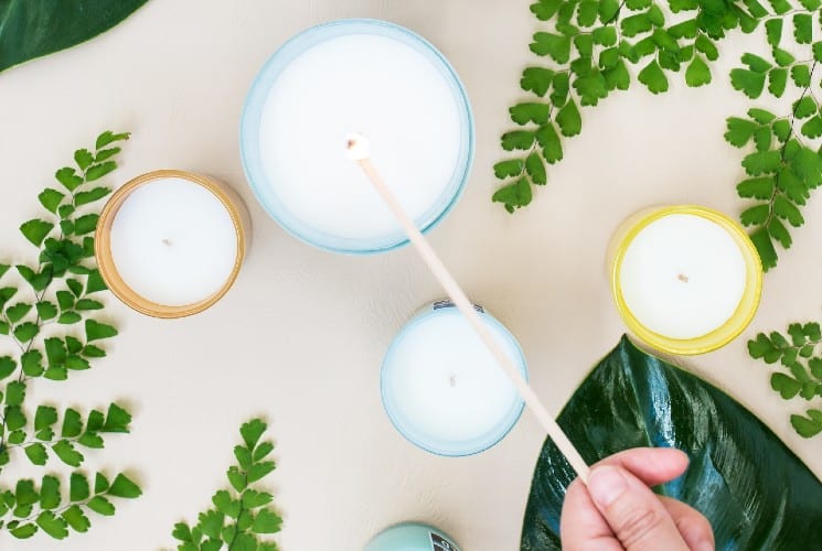 Several candles on a table with green leaves and a person lighting one candle with a long wooden stick