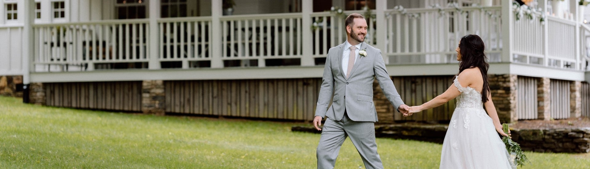 A groom in a grey suit holding the hand of his bride in white walking across the lawn in front of a large white home