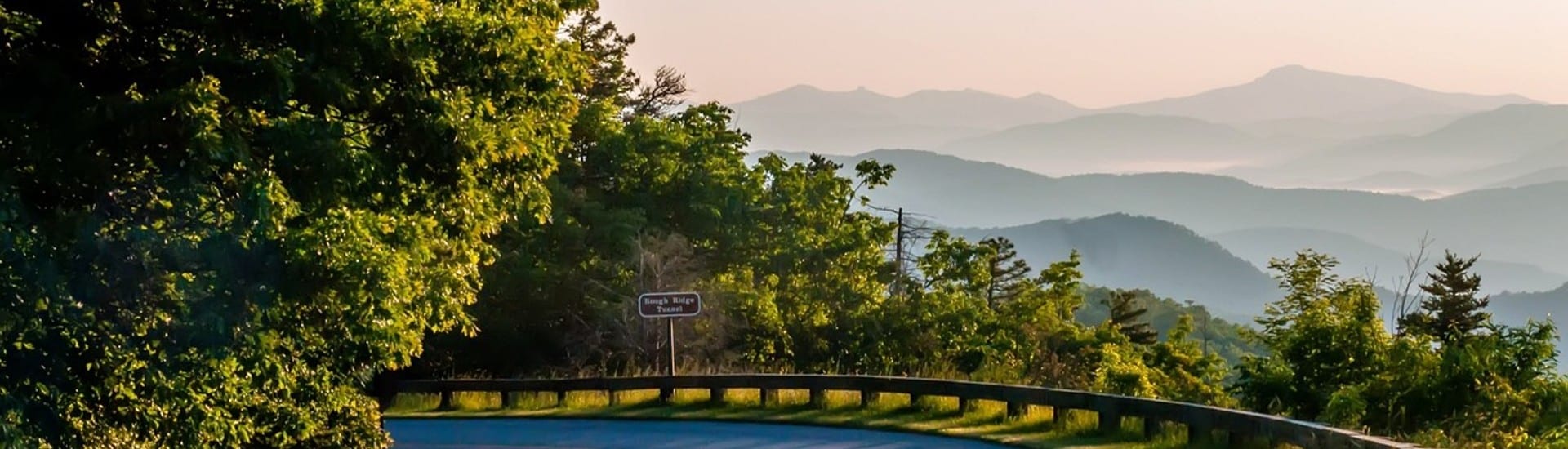 A curved road at the edge of an expansive view of rolling mountains covered in trees in a hazy mist