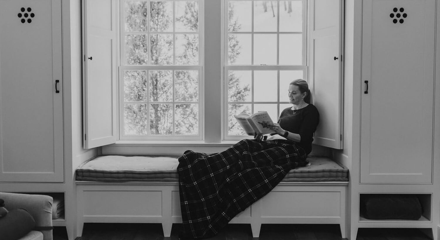 A woman under a blanket reading a book on a bench below a large window with shutters