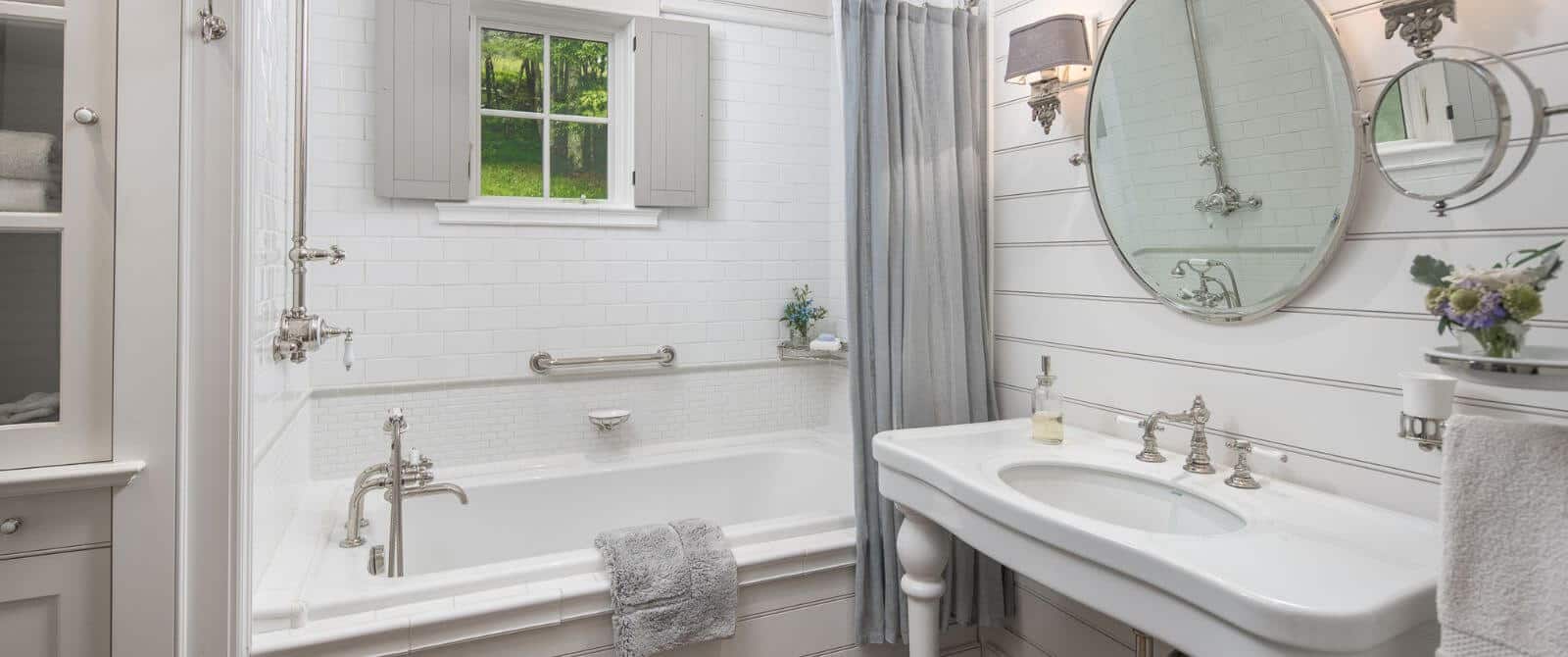 Elegant white bathroom with shiplap walls, pedestal sink, round mirror and shower with tub and grey curtain