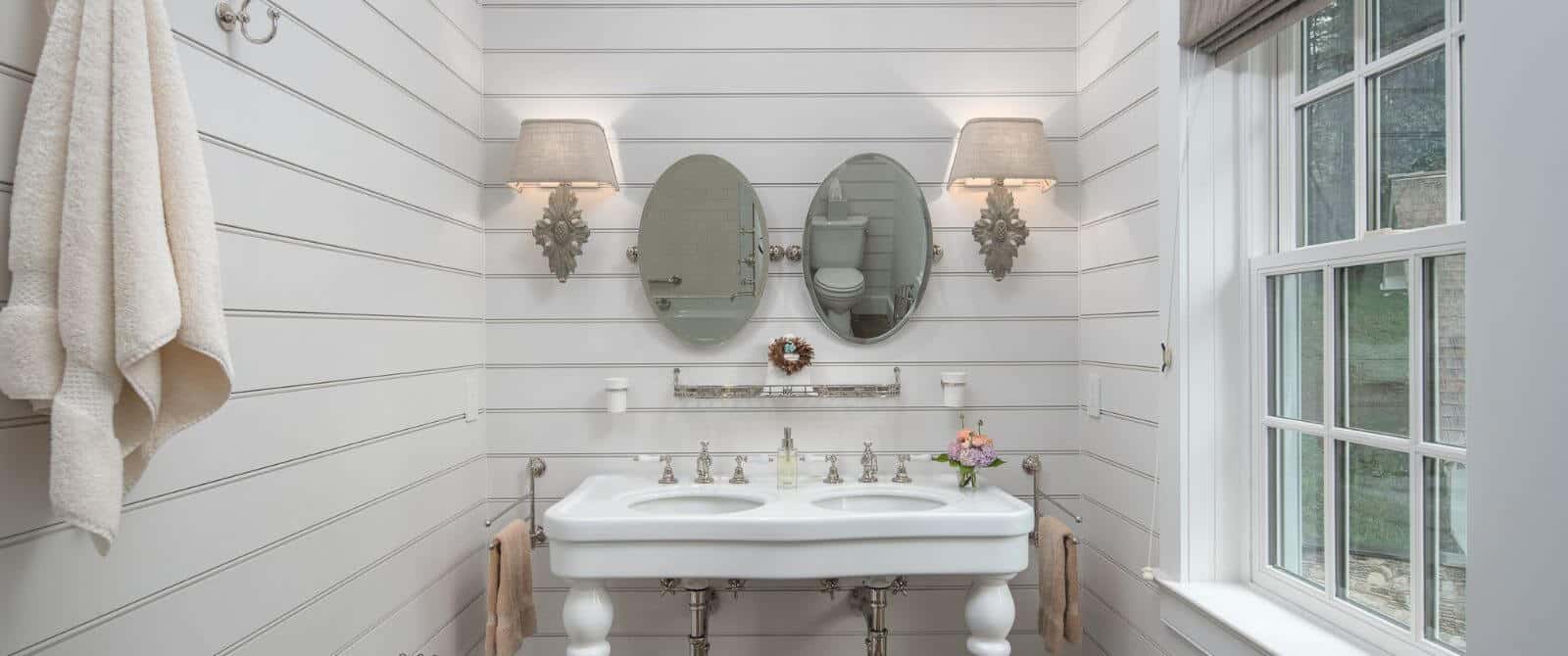 Bathroom with double bowl pedestal sink, two oval mirrors, lamp sconces and a bright window