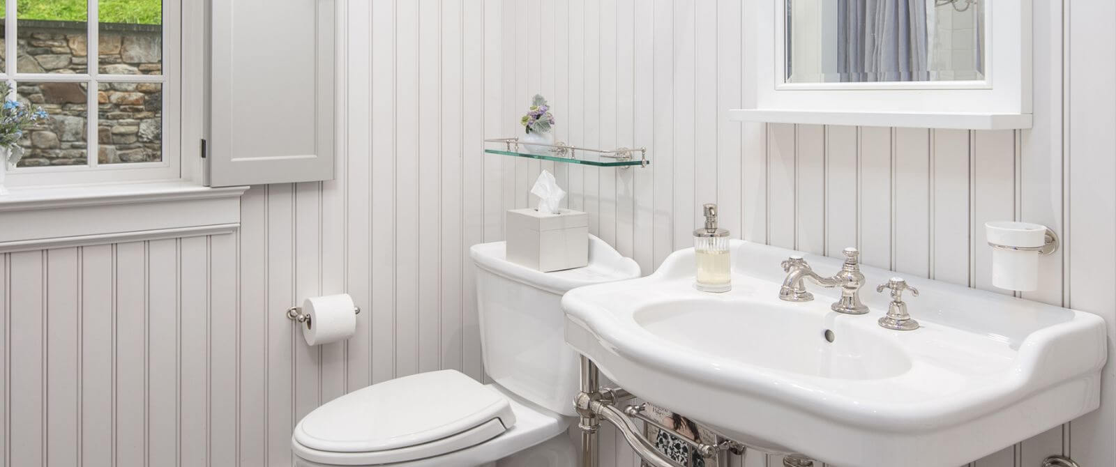White bathroom with pedestal sink, framed mirror and toilet by a window