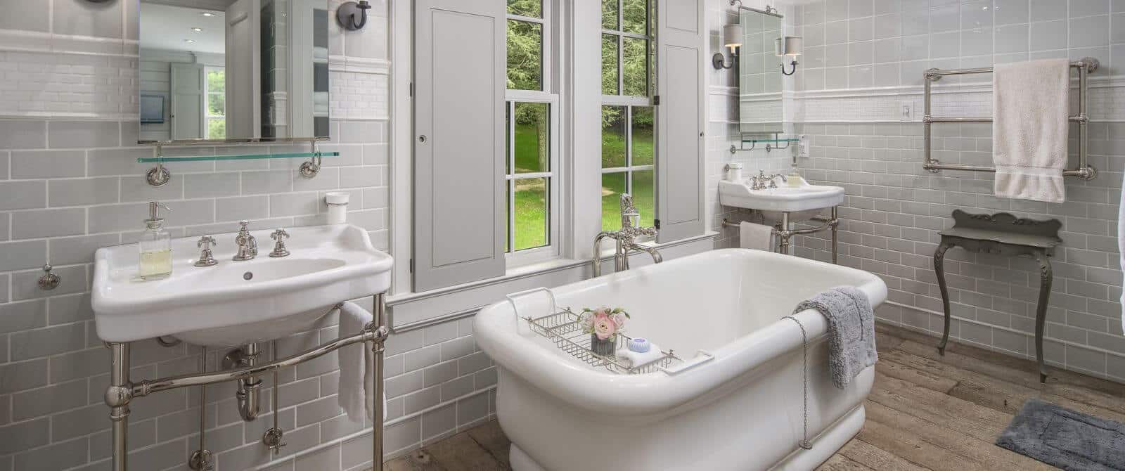 Bathroom with two pedestal sinks, each with a large mirror and soaker tub below a large window overlooking the outdoors