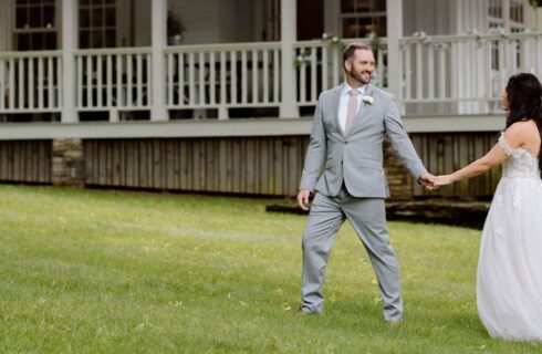 A groom in a grey suit holding the hand of his bride in white walking across the lawn in front of a large white home