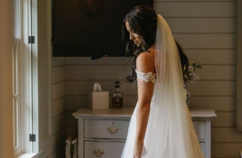 A bride in her white dress and veil standing in a bedroom in front of a bright window