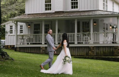 A groom in a grey suit holding the hand of his bride in white walking across the lawn in front of a large white home