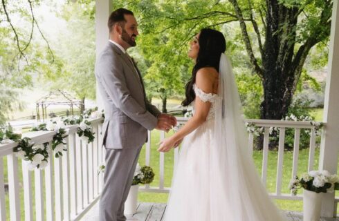 A bride and groom standing on an outdoor porch with greens and white flowers draped around the railing