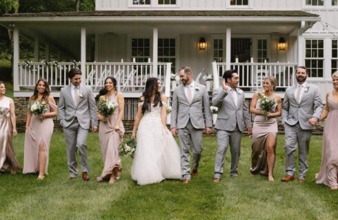 A bride and groom and their wedding party walking in a line on a lawn on front of a large white home