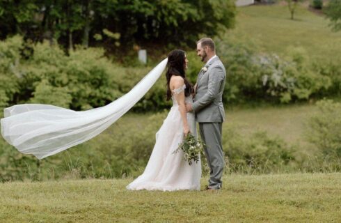 A bride with her veil blowing in the wind standing with a groom in a field surrounded by trees