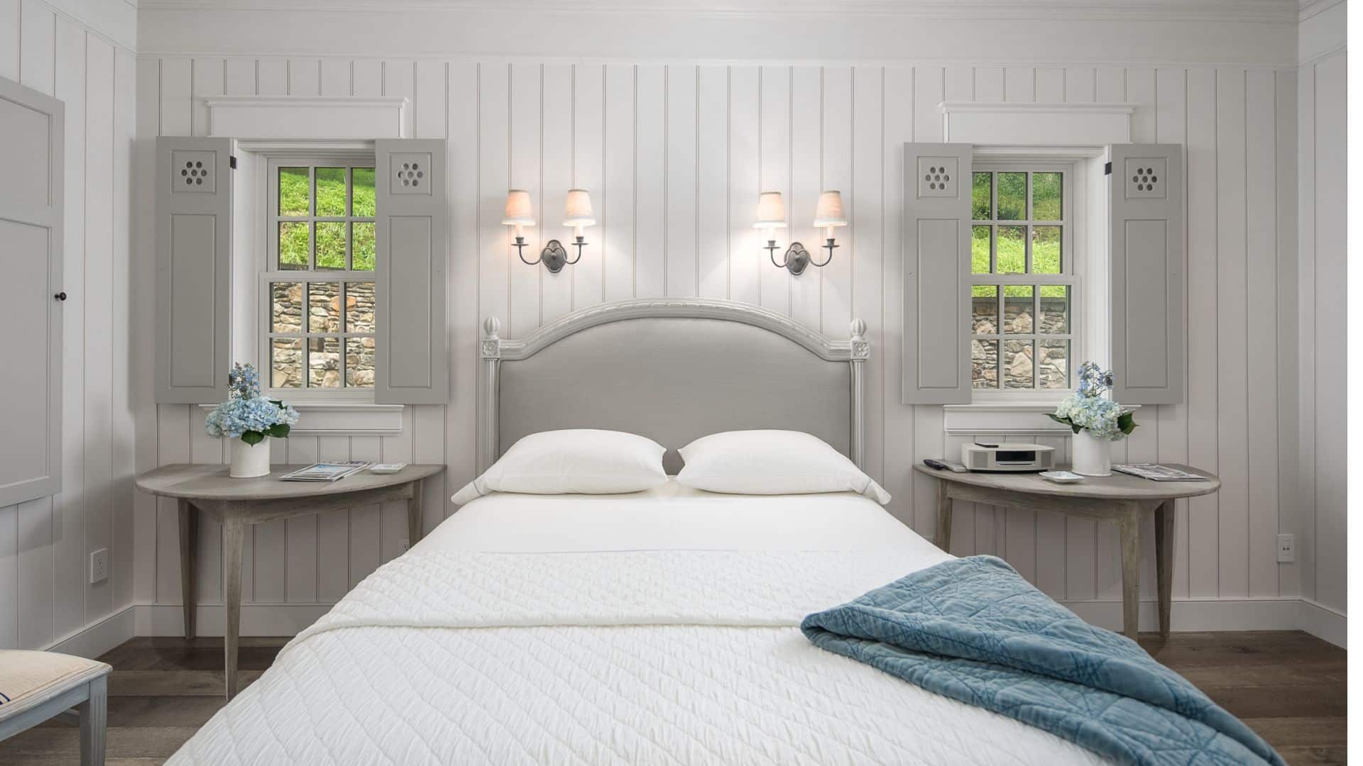 Bright and elegant bedroom with a queen bed, two windows with shutters and side tables with vases with blue flowers