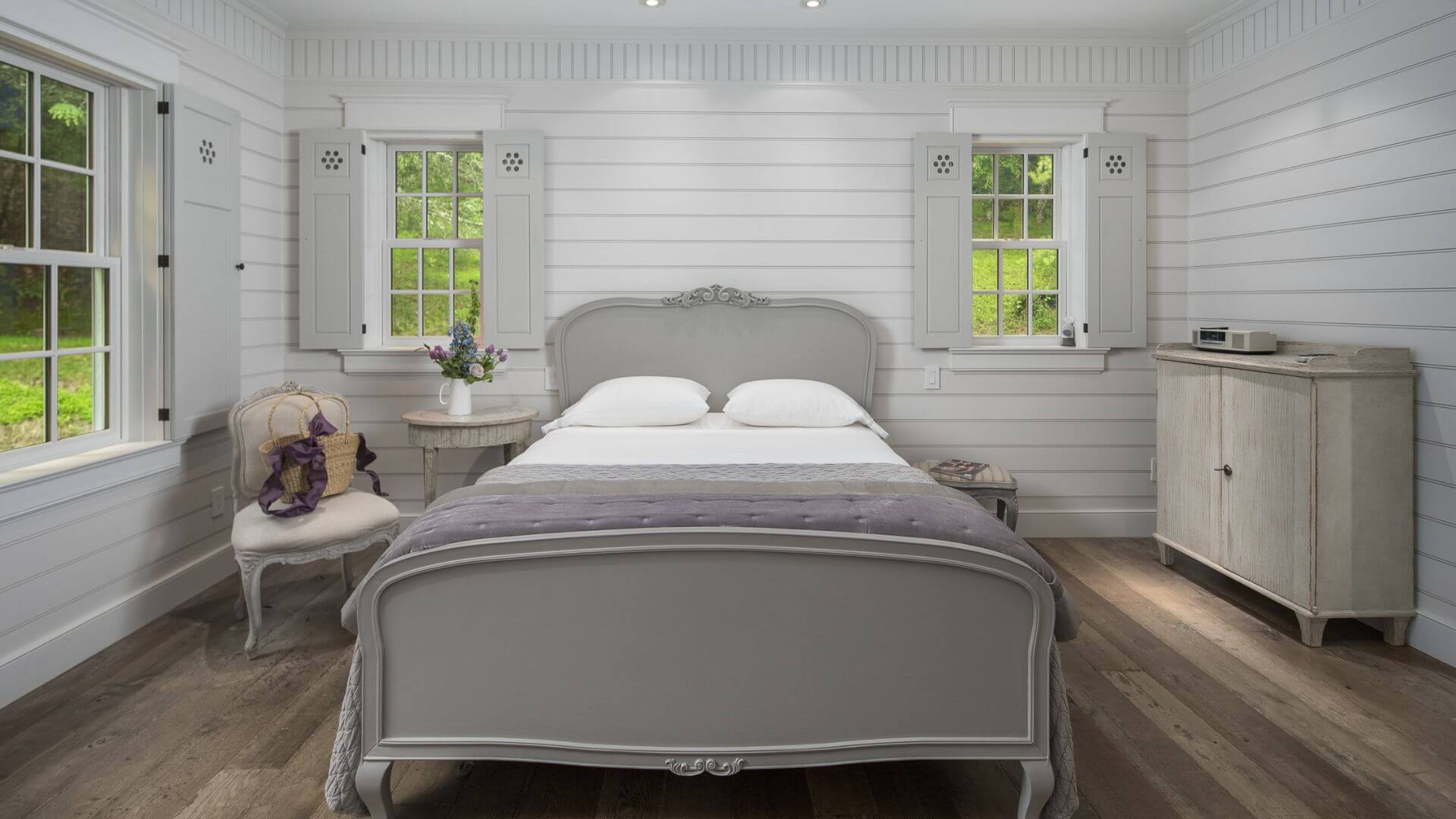 Elegant bedroom with a queen bed, dresser, side table with vase of flowers, chair and bright windows