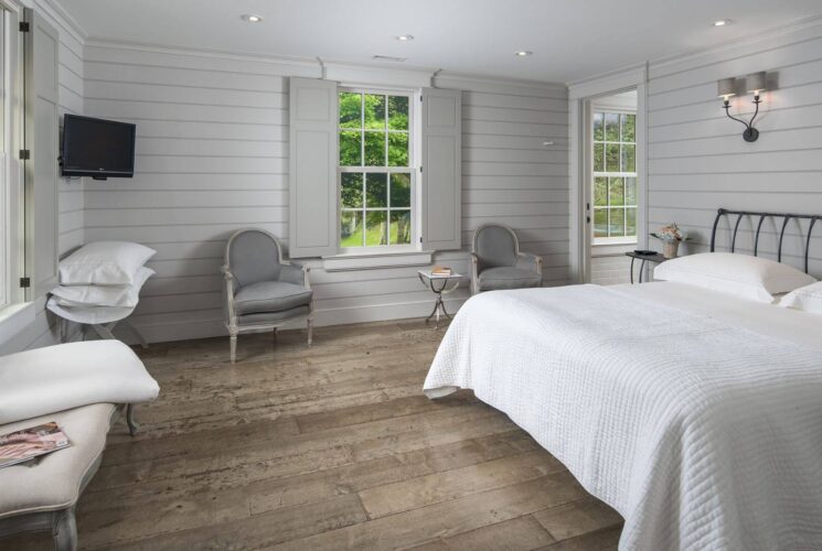 Large bedroom with shiplap walls, sitting chairs, TV, large windows and doorway into a bathroom