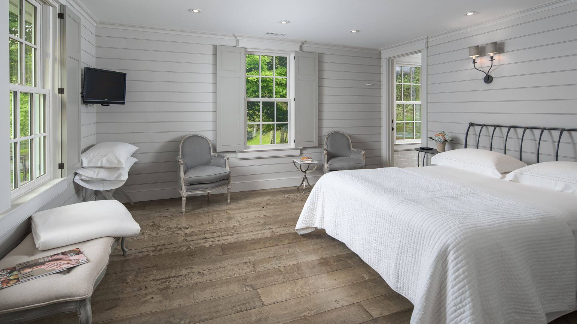 Large bedroom with shiplap walls, sitting chairs, TV, large windows and doorway into a bathroom