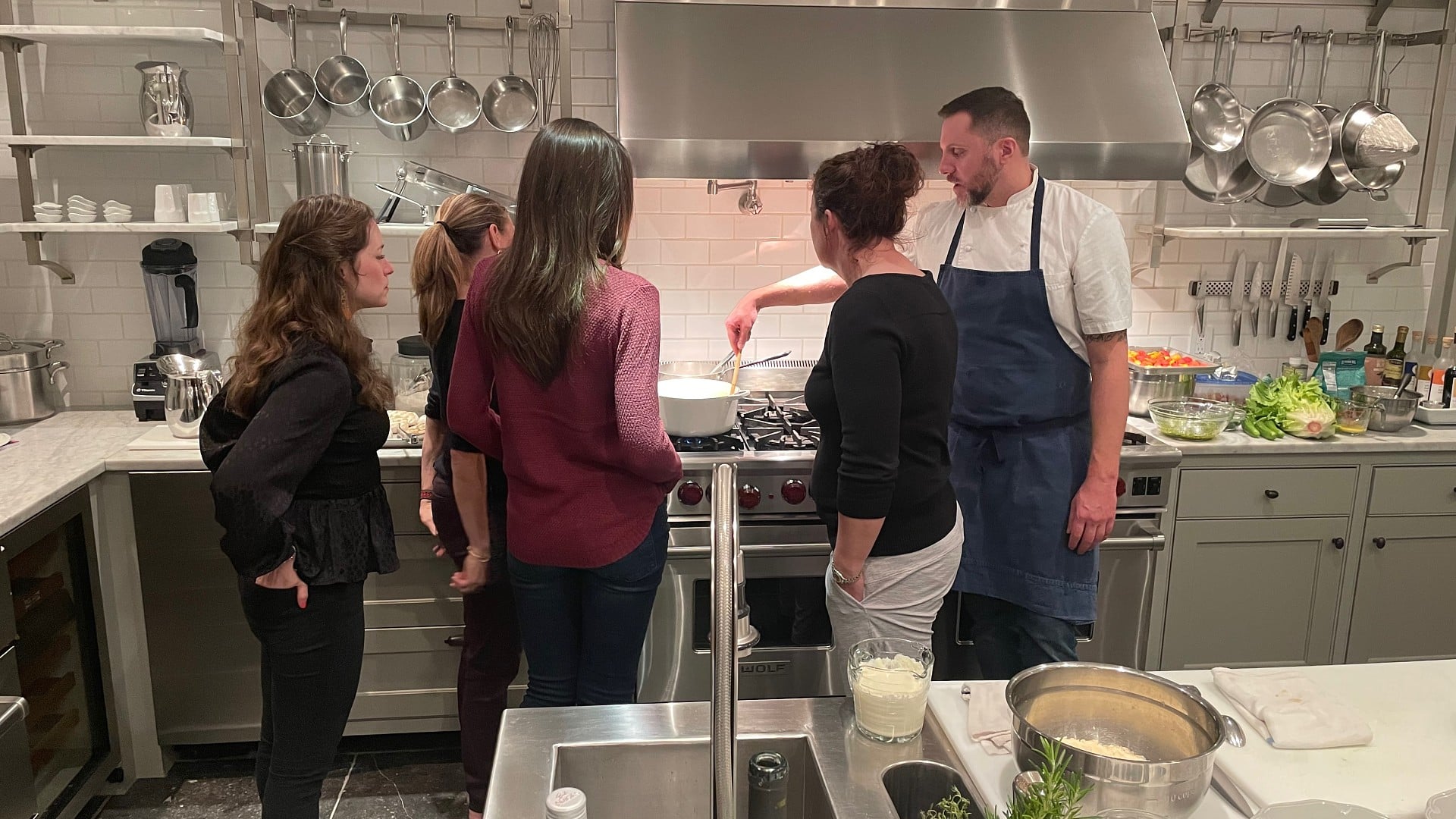 A chef in a blue apron standing at the stove in a chef's kitchen teaching four women