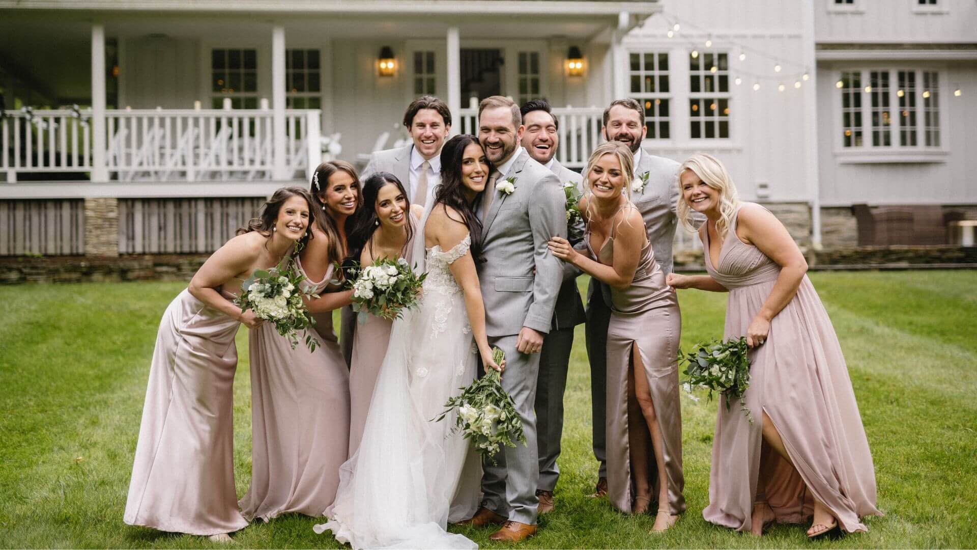 A bride and groom and their wedding party huddled together for a photo on the lawn in front of a large white home