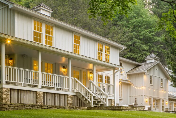 Front facade of a large two-story white home with wrap around porch, string lights and surrounded by tall trees