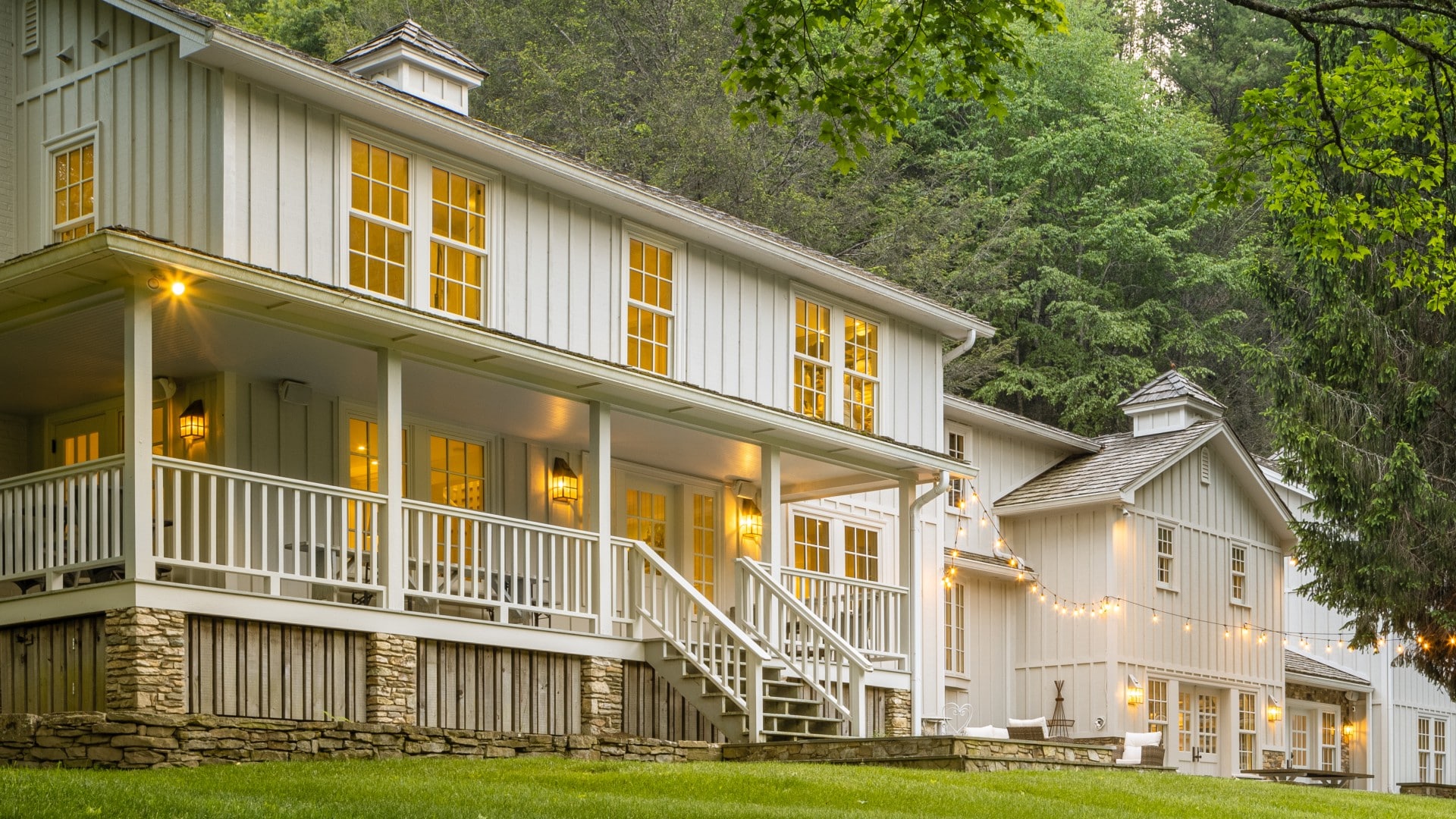 Front facade of a large two-story white home with wrap around porch, string lights and surrounded by tall trees