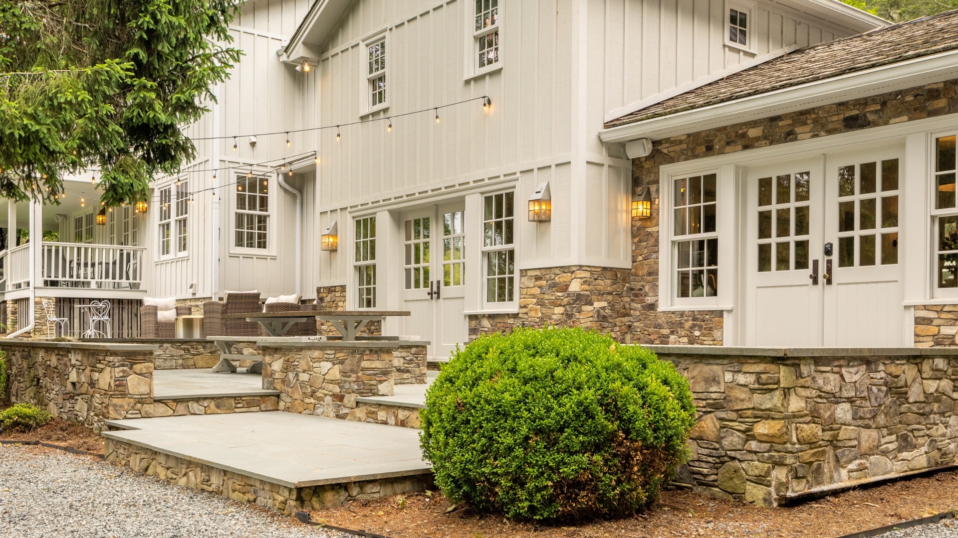 Expansive outdoor stone patio of a white home with picnic table, string lights and green bushes