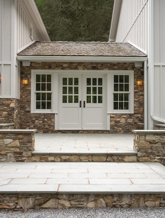 A portico walkway between two large areas of a white home with an expansive outdoor stone patio