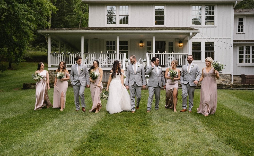 A bride and groom and their wedding party walking in a line on a lawn on front of a large white home