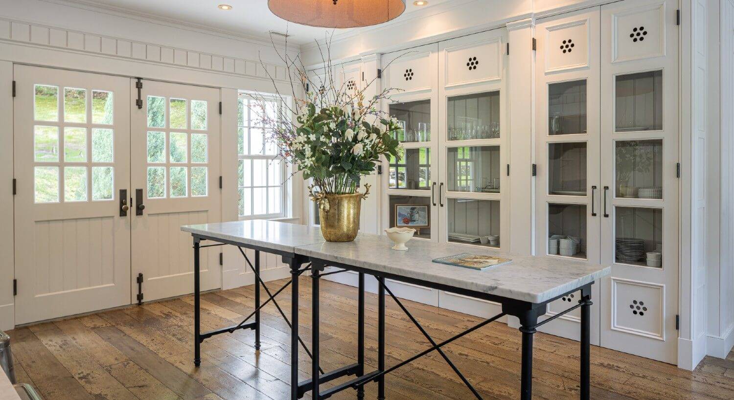Entry area of a home with built in cabinets with glass doors, hardwood floors and long marble table with gold vase of flowers