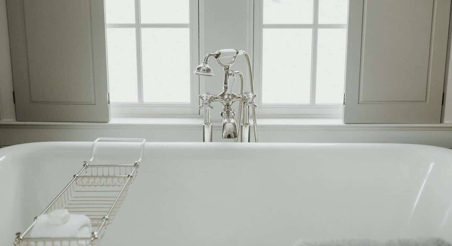 Large white soaker tub with elegant silver details in front of a bright window with shutters